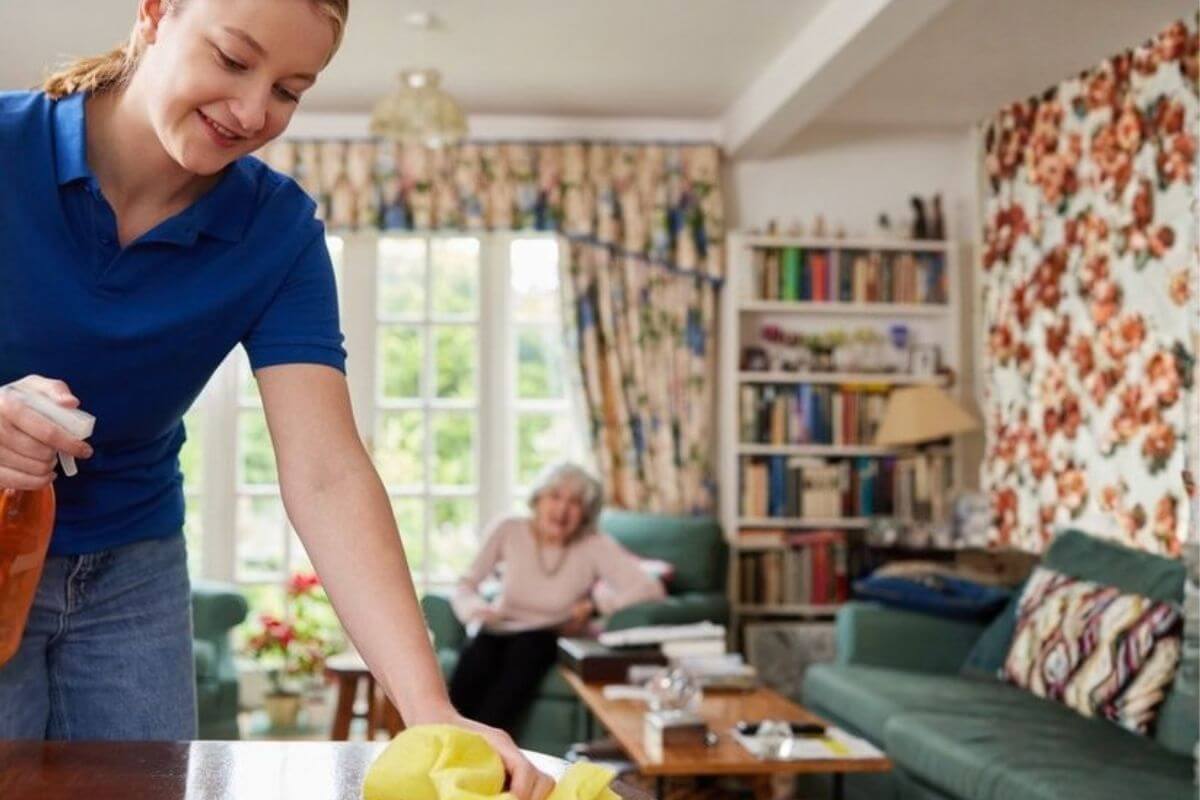 Haushaltshilfe reinigt einen Tisch im Wohnzimmer, während eine Seniorin auf dem Sofa sitzt.