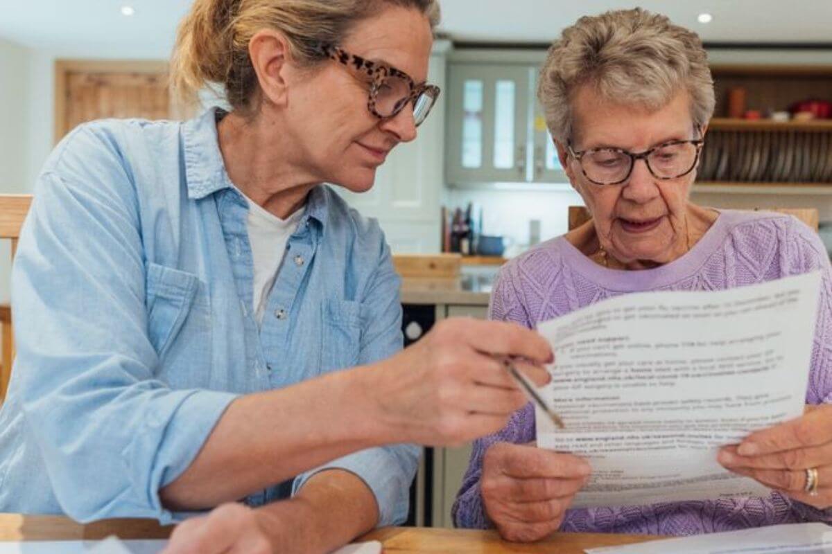 Alltagshelferin unterstützt eine Seniorin beim Lesen eines Dokuments am Tisch.